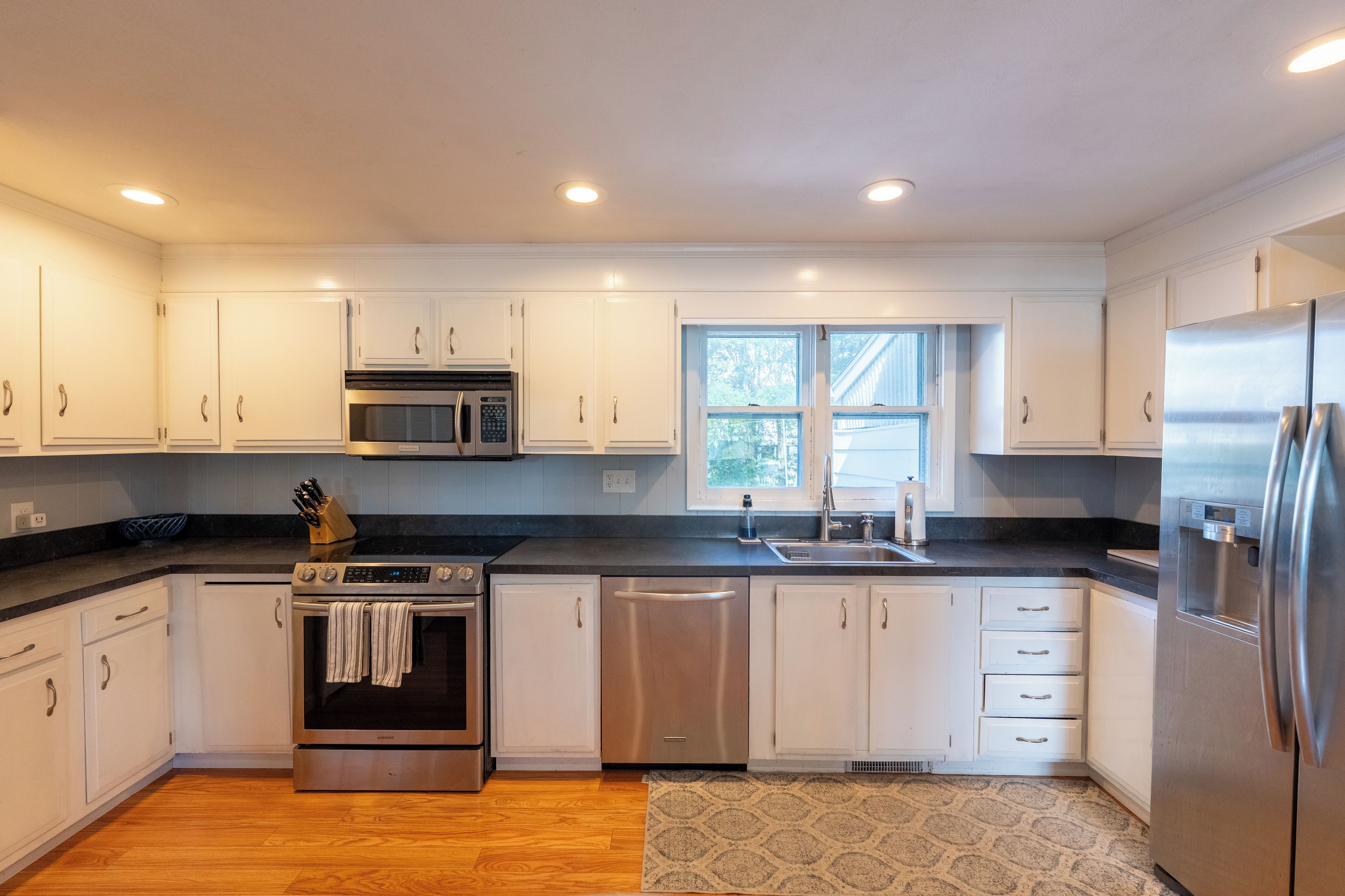 A modern kitchen with white cabinets, stainless steel appliances, and a window providing natural light.