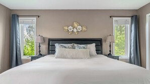 Ein großes Bett mit dunklem Kopfteil, an dessen Seiten jeweils ein Nachttisch mit Lampe steht, in einem Zimmer mit beige gefärbten Wänden und einer Dekoration an der Wand über dem Bett.