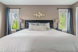 A large bed with a dark headboard, flanked by two bedside tables with lamps, in a room with beige walls and a decorative wall piece above the bed. - Cottage Cove, 7Beds, Family Retreat by Sylvan Lake (Sylvan Lake)