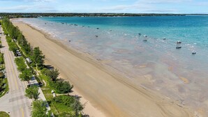 Ein Strand mit klarem Wasser, einer parallel zur Küste verlaufenden Straße und einer entfernten Küstenlinie mit Gebäuden.