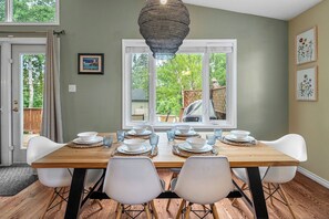 A dining area with a wooden table set for four, white chairs, and a hanging light fixture. - Cottage Cove, 7Beds, Family Retreat by Sylvan Lake (Sylvan Lake)