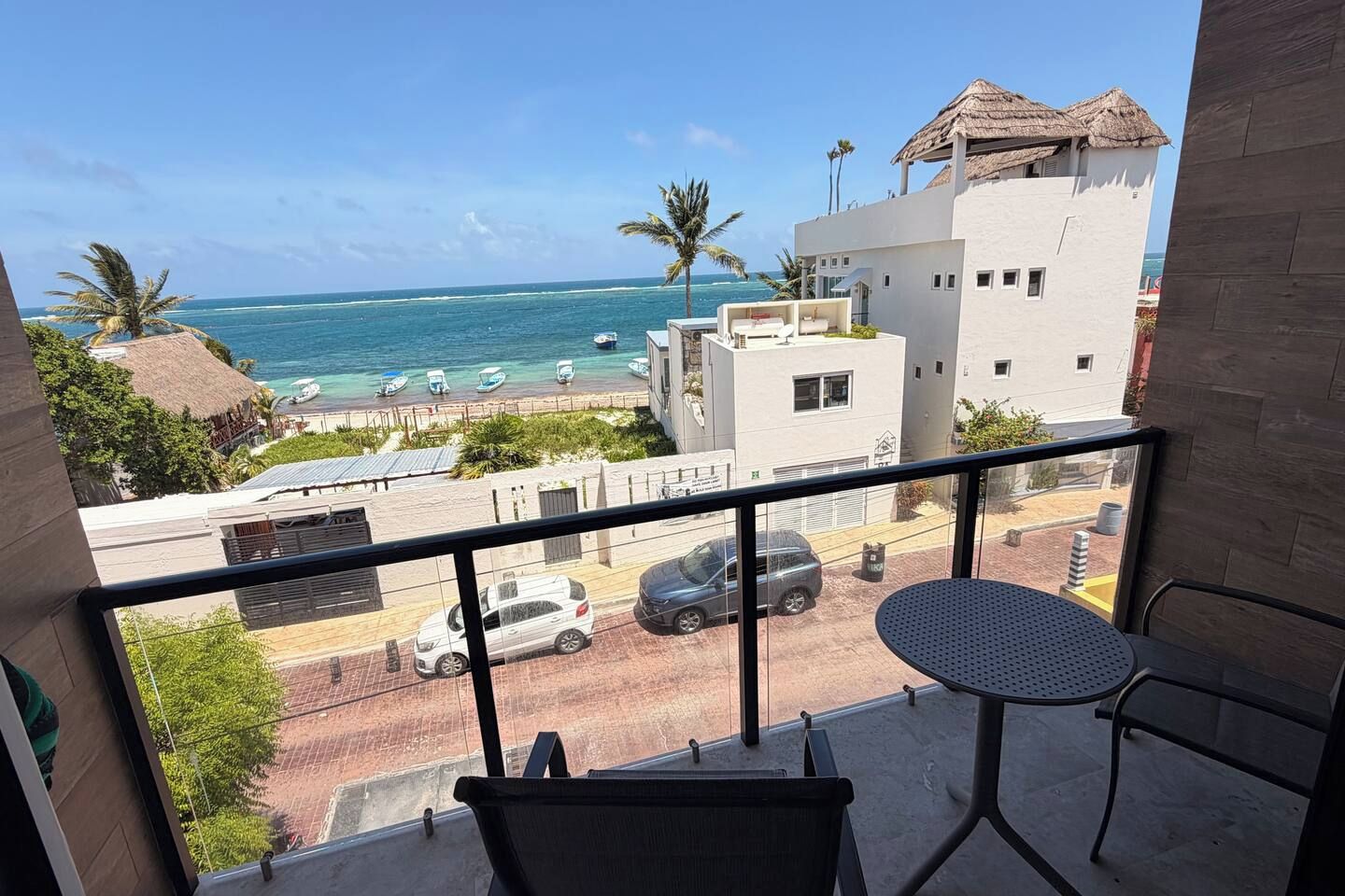 A balcony with a view of the ocean, a street with cars, and a building with a thatched roof.