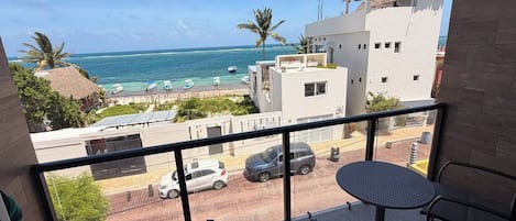 A balcony with a view of the ocean, a street with cars, and a building with a thatched roof.