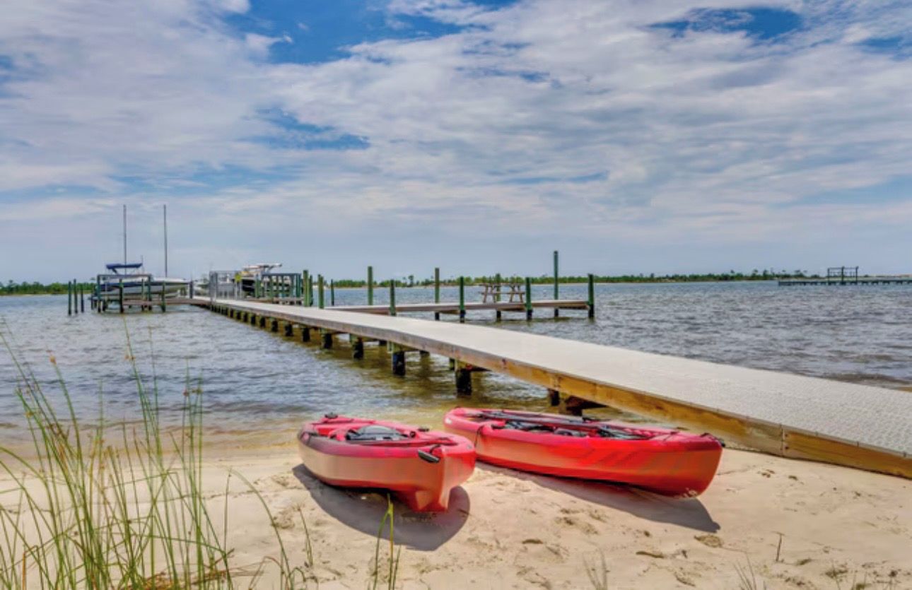 A pier extending into calm waters with kayaks on the sand.