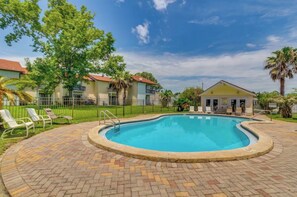 A swimming pool with a brick deck, surrounded by lounge chairs and a fenced area with buildings in the background.