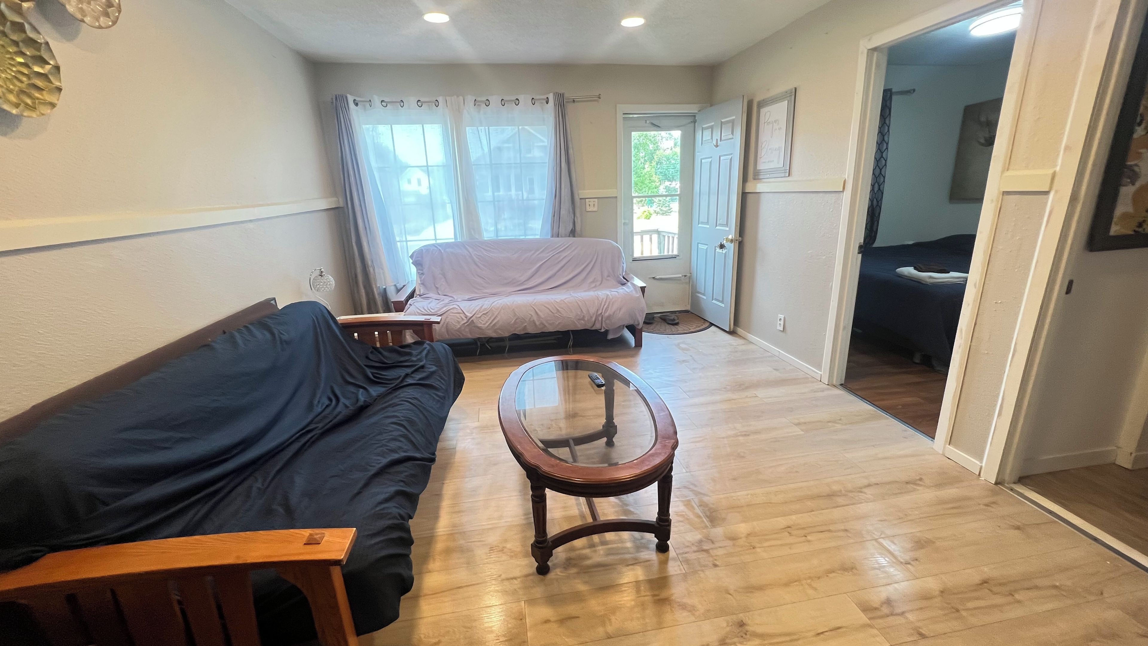 A room with a wooden floor, a sofa, a glass-top coffee table, a bed with a white cover, and a door leading to another room.