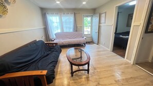 A room with a wooden floor, a sofa, a glass-top coffee table, a bed with a white cover, and a door leading to another room. - Cozy 2-bedroom house in brilliant Dilworth with AC (Dilworth)
