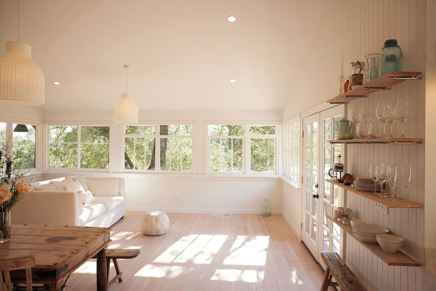 Une pièce lumineuse, baignée de soleil, avec un canapé blanc, une table en bois et des étagères remplies de verres.