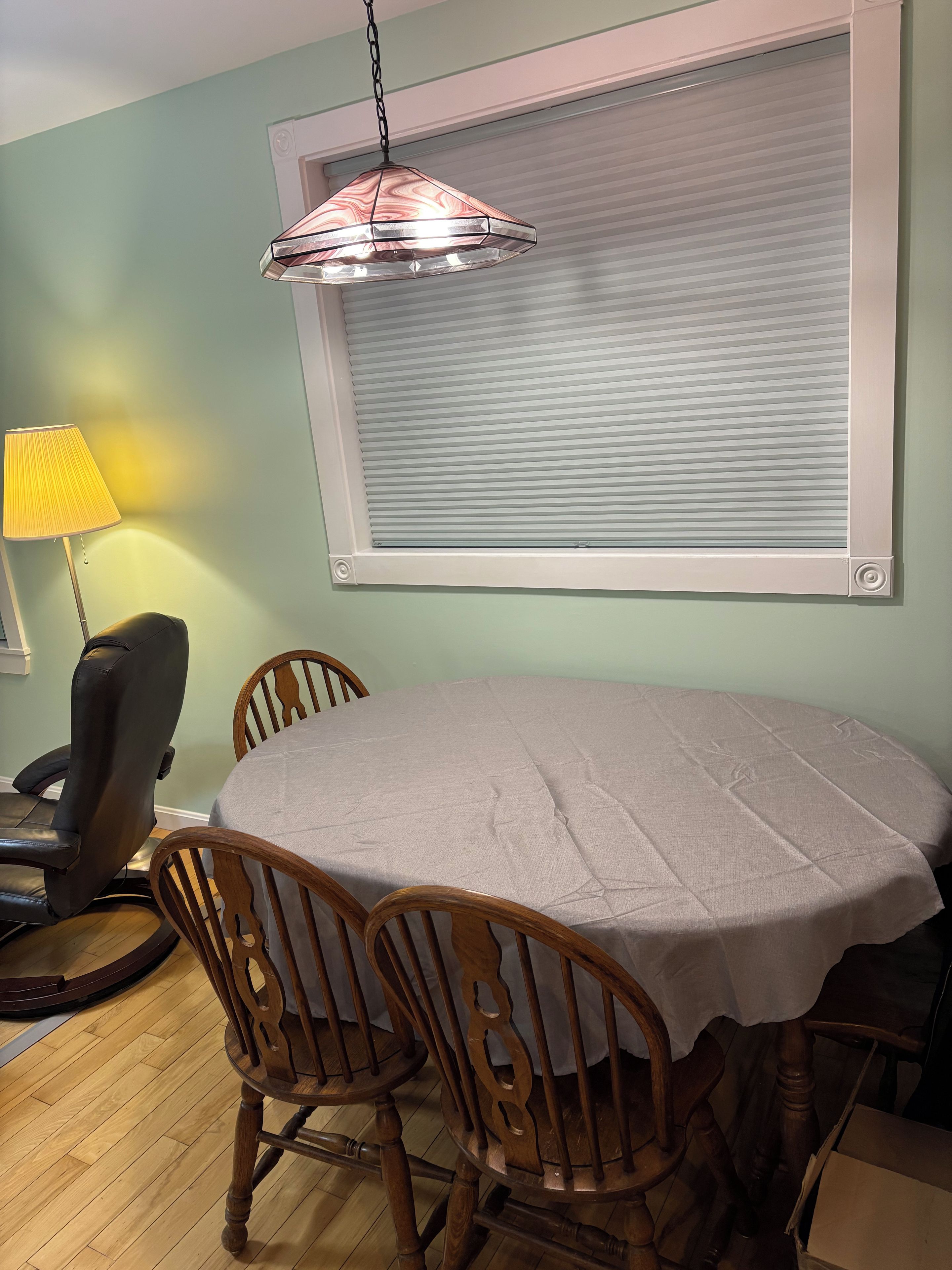 A dining area with a round table, four wooden chairs, a lamp, and a window with blinds.