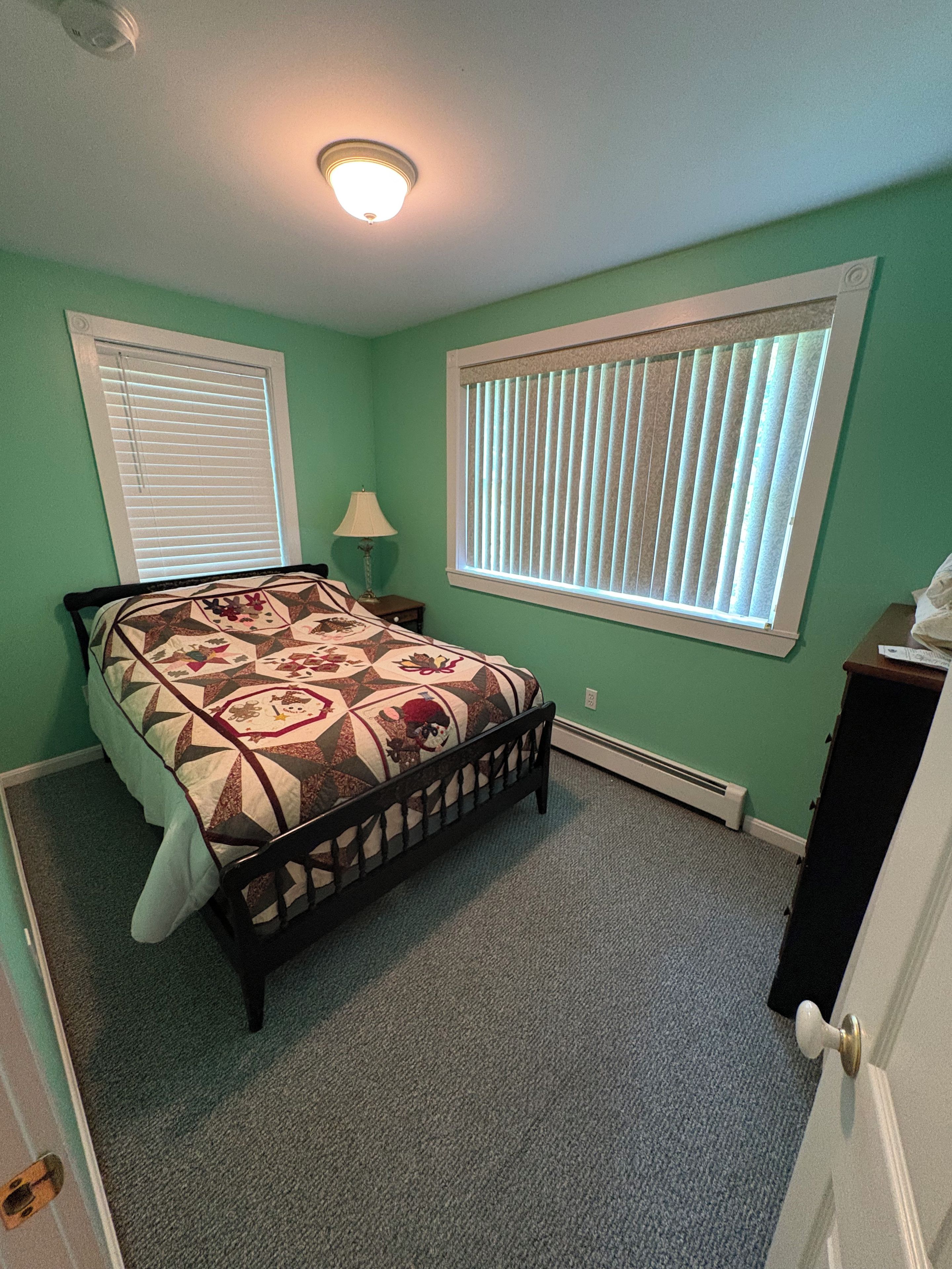 A bedroom with a bed, a nightstand, a lamp, a window with blinds, and a door.