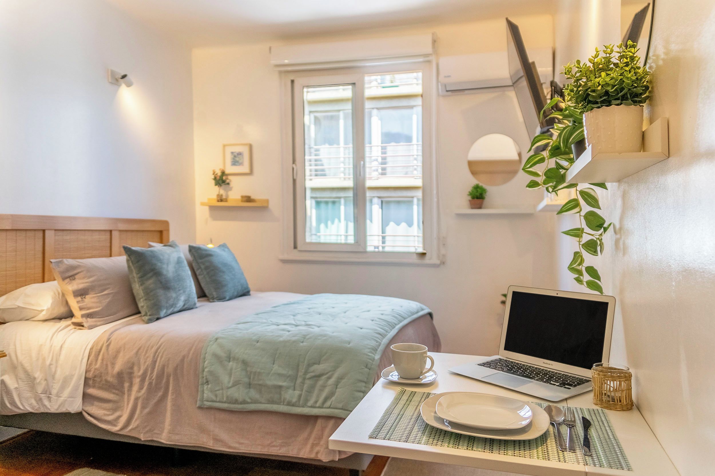Un dormitorio con una cama, un ordenador portátil sobre una mesa, una taza y una planta.