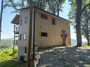 A two-story cabin with a balcony, a gravel driveway, and a view of the surrounding forest and hills. - Appalachian Mountain Retreat (Boone)