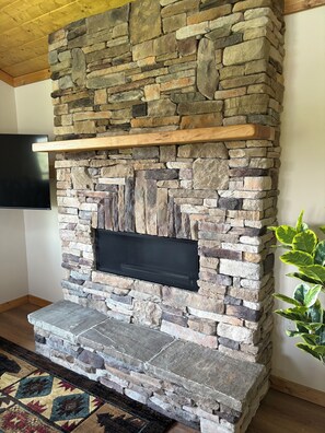 A stone fireplace with a flat-screen TV above it, a wooden shelf, and a patterned rug on the floor. - Appalachian Mountain Retreat (Boone)