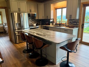A kitchen with a marble countertop, stainless steel appliances, and wooden bar stools. - Appalachian Mountain Retreat (Boone)