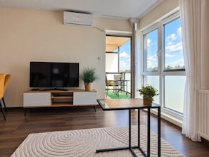 A modern living room with a flat-screen TV, a sofa, a coffee table, and a balcony with a view.