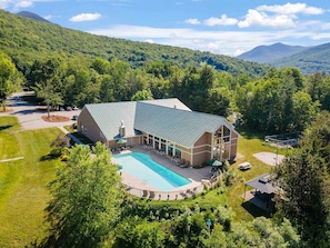 A large building with a swimming pool, surrounded by lush greenery and mountains in the background. - New 2026 Build · 3 Min to Loon · Movie Room · Pool & Hot Tub · Sleeps 20 (Lincoln)