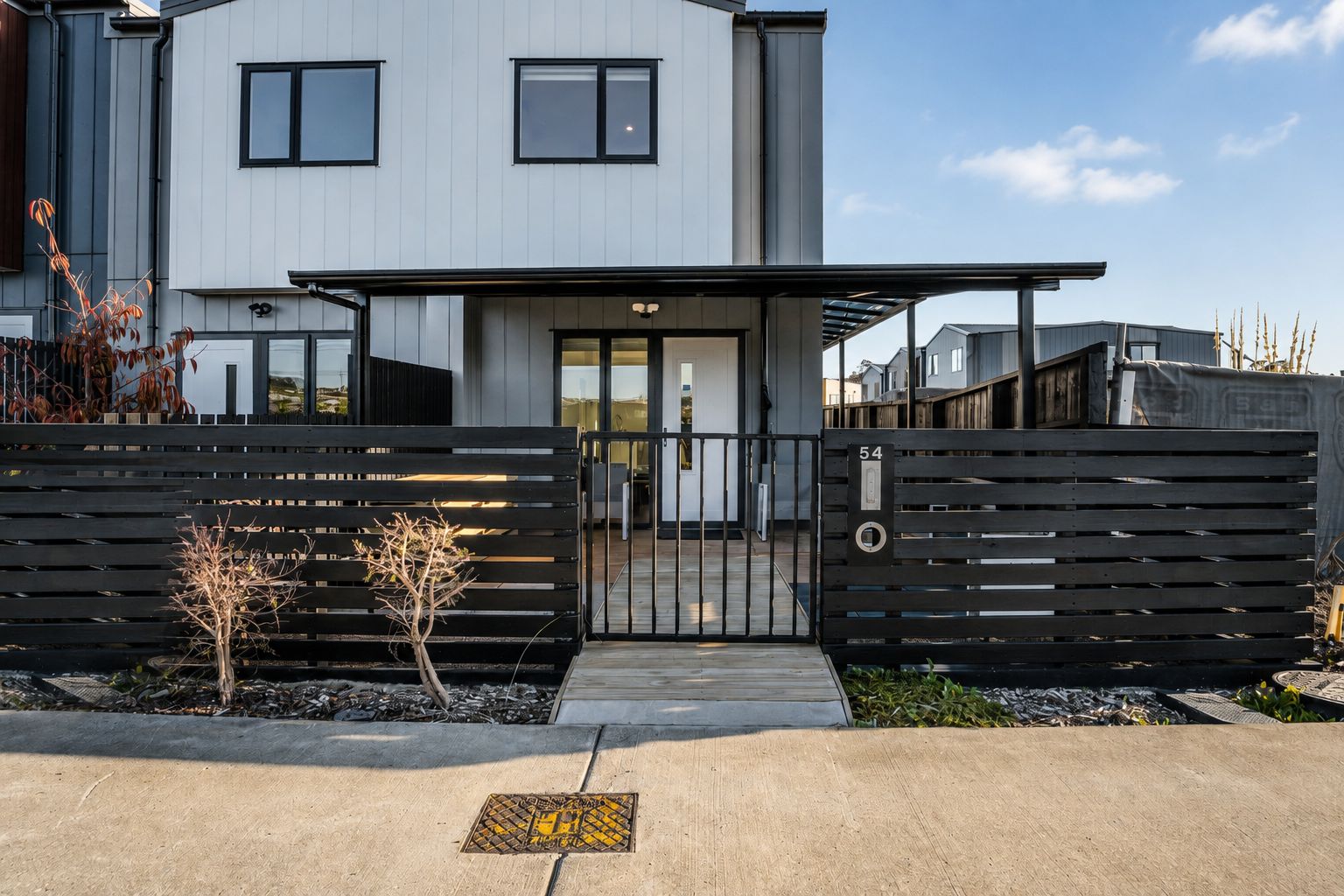 A modern two-story house with a gated entrance, a small garden with plants, and a clear sky.