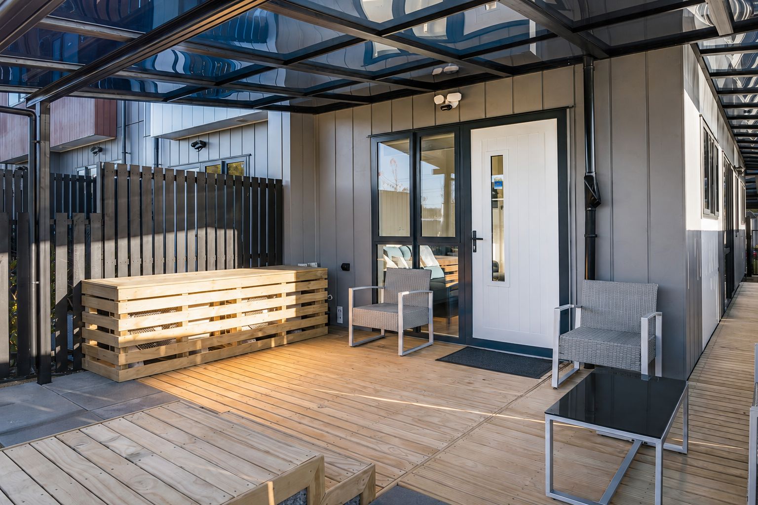 A covered outdoor seating area with wooden benches and chairs, a glass door leading inside, and a wooden deck.
