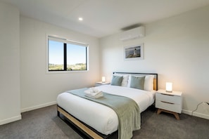 A bedroom with a bed, bedside tables, a window, an air conditioning unit, and a painting on the wall. - Modern & New 3BR - 2.5 bath Haven Near Totara Park (Totara Park)