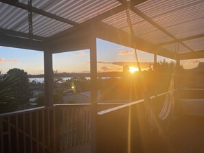 A covered outdoor area with a hammock, a view of the sunset over a body of water, and a distant landscape. - Pukehina beach house: Estuary views + Outdoor bath (Pukehina)