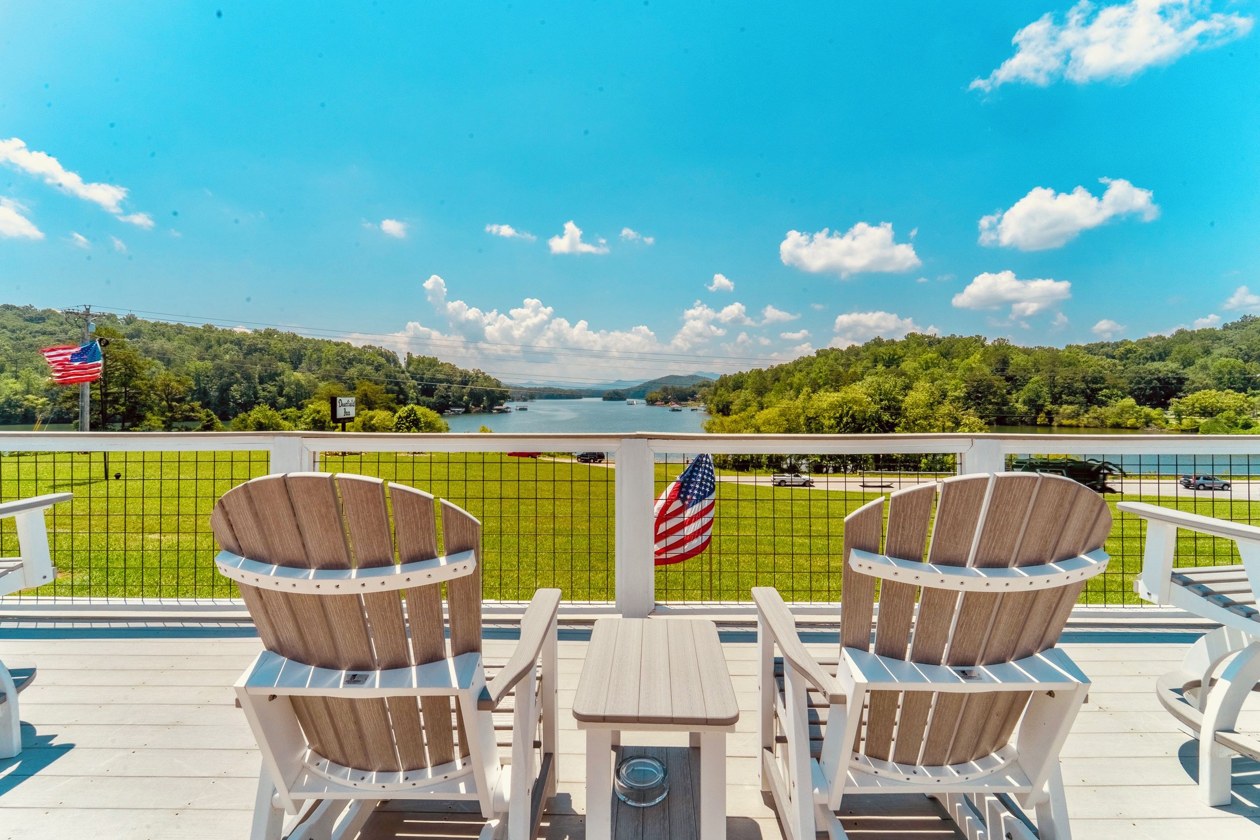 A deck with two wooden chairs and a table, overlooking a lake with a view of a forested area and a distant body of water.