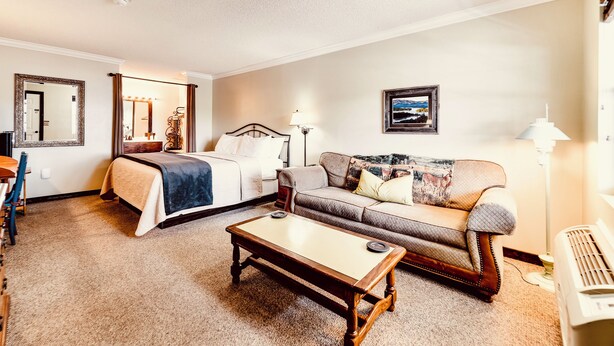 A hotel room with a bed, a sofa, a coffee table, a desk, and a lamp. - Deerfield Inn - King Bed w/ Sofa (Hayesville)