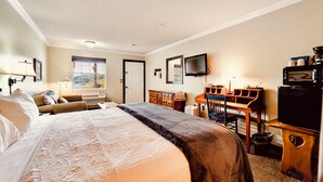 A hotel room with a bed, a desk, a chair, a television, and a window with a view. - Deerfield Inn - King Bed w/ Sofa (Hayesville)