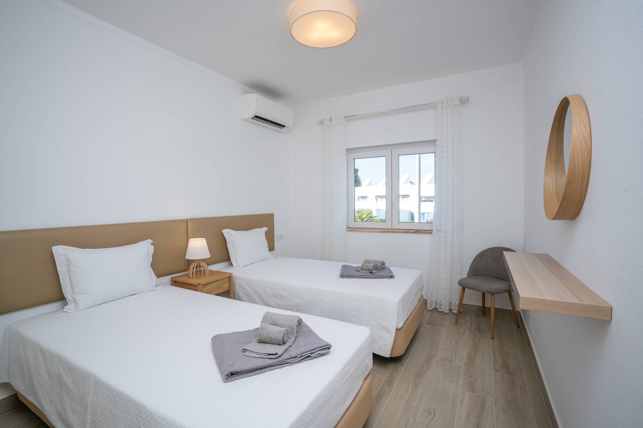 Habitació d'hotel amb dos llits, una escriptori, una cadira, una finestra amb cortines i un mirall fixat a la paret.