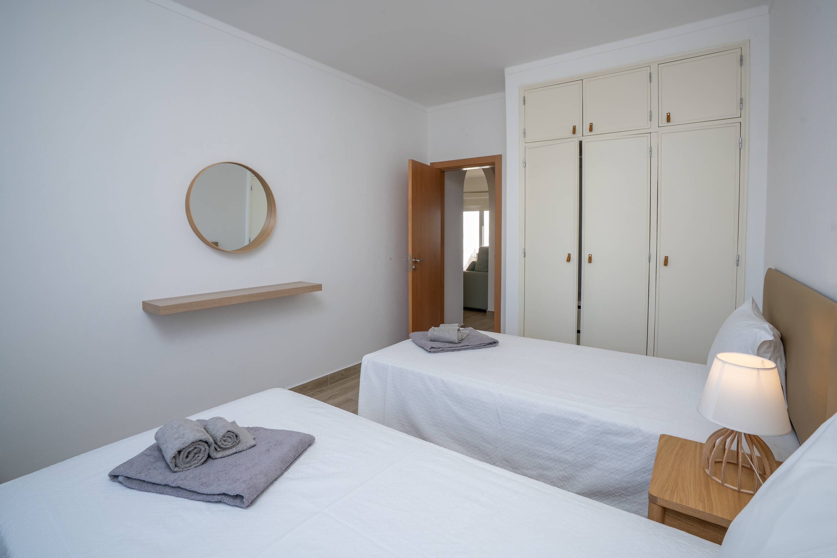 A hotel room with two single beds, a wooden nightstand with a lamp, a mirror on the wall, and a wardrobe with multiple doors.