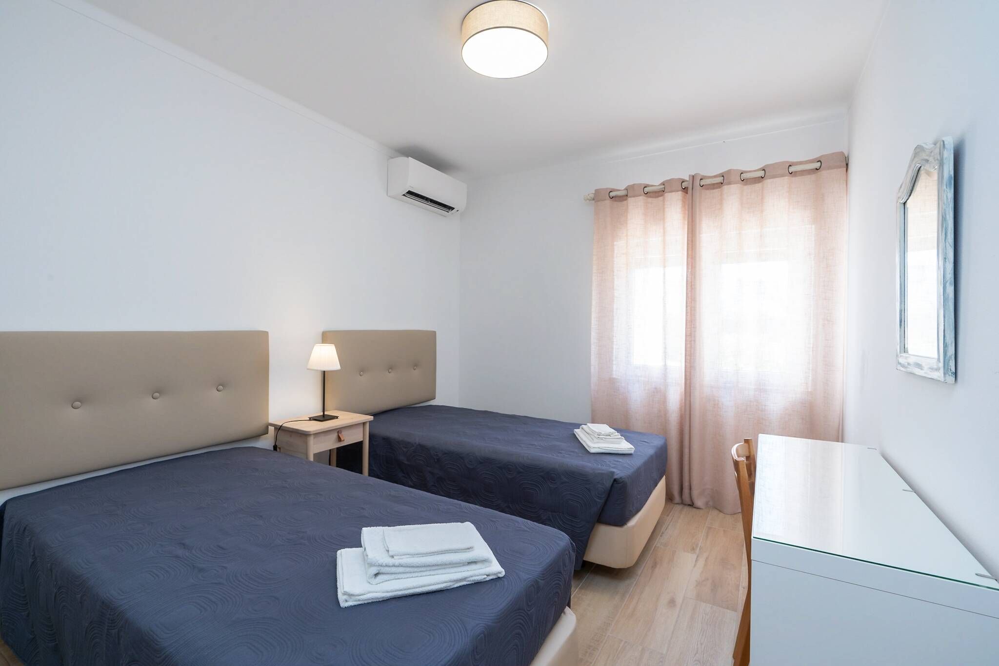 Une chambre d’hôtel avec deux lits, une table de chevet avec une lampe, un bureau et une fenêtre avec des rideaux.
