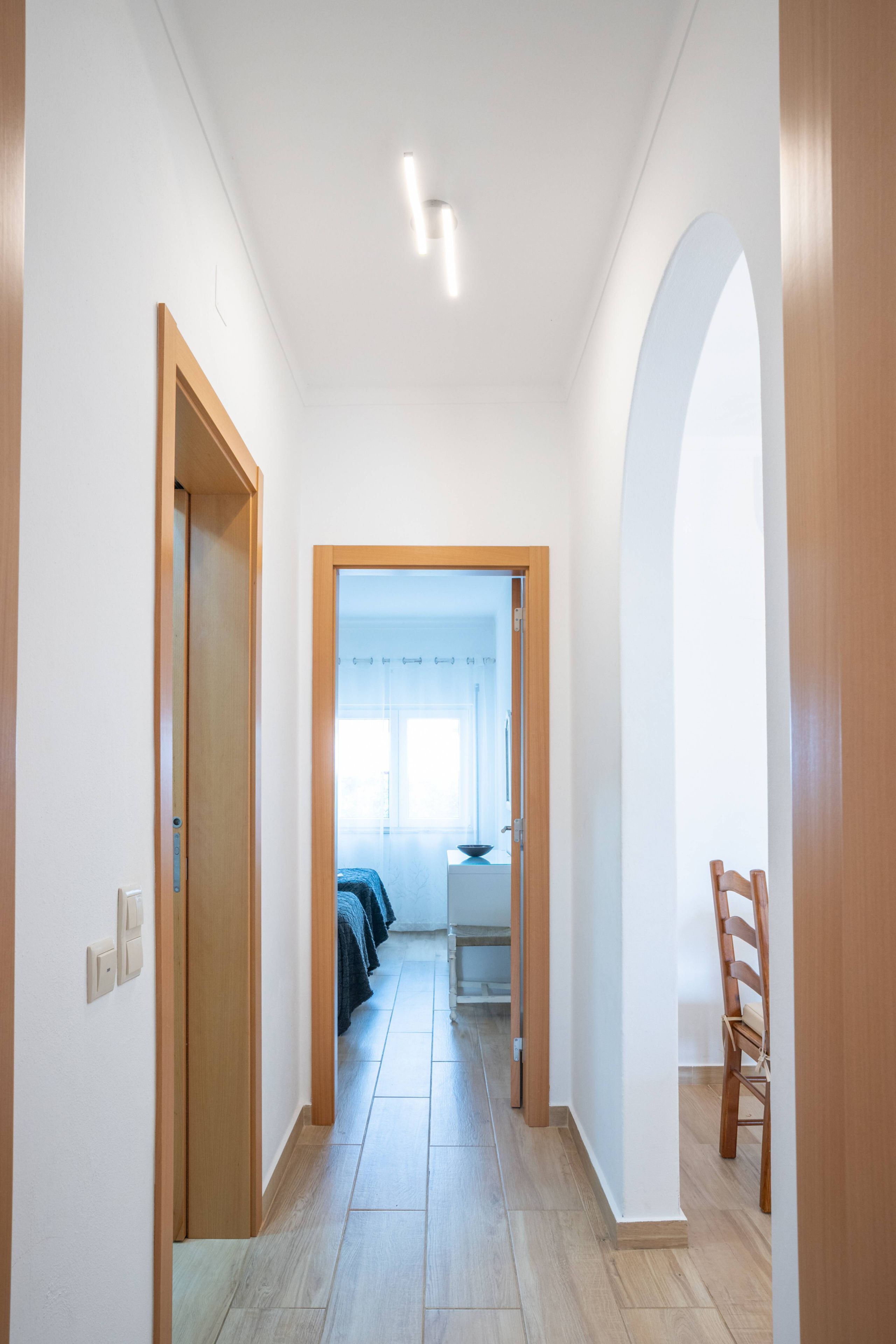 Un couloir avec des portes en bois, un banc et une vue sur une pièce où se trouvent un lit et une fenêtre.