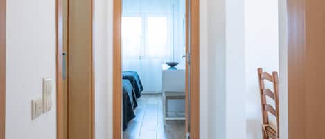 Un couloir avec des portes en bois, un banc et une vue sur une pièce où se trouvent un lit et une fenêtre.
