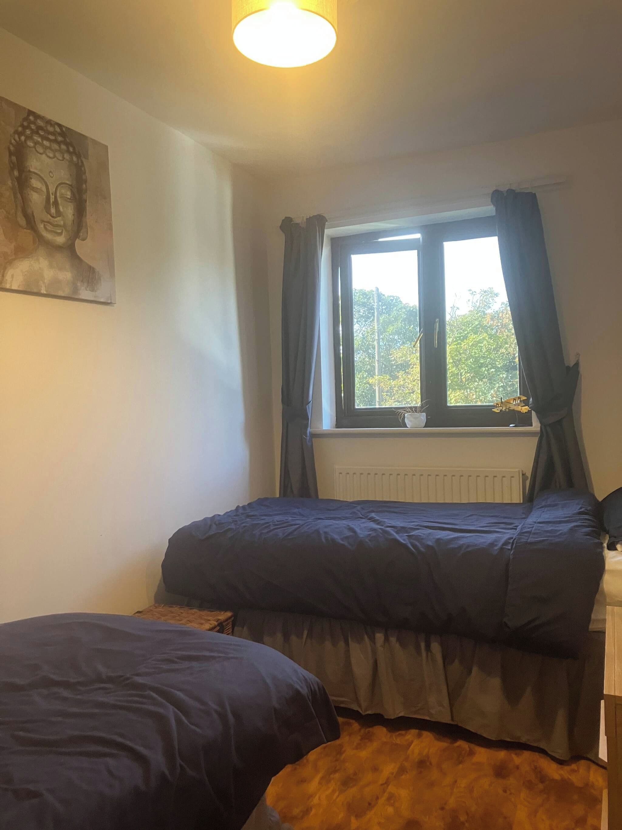 Quarto com duas camas, janela com cortinas, radiador e estátua de Buda na parede.