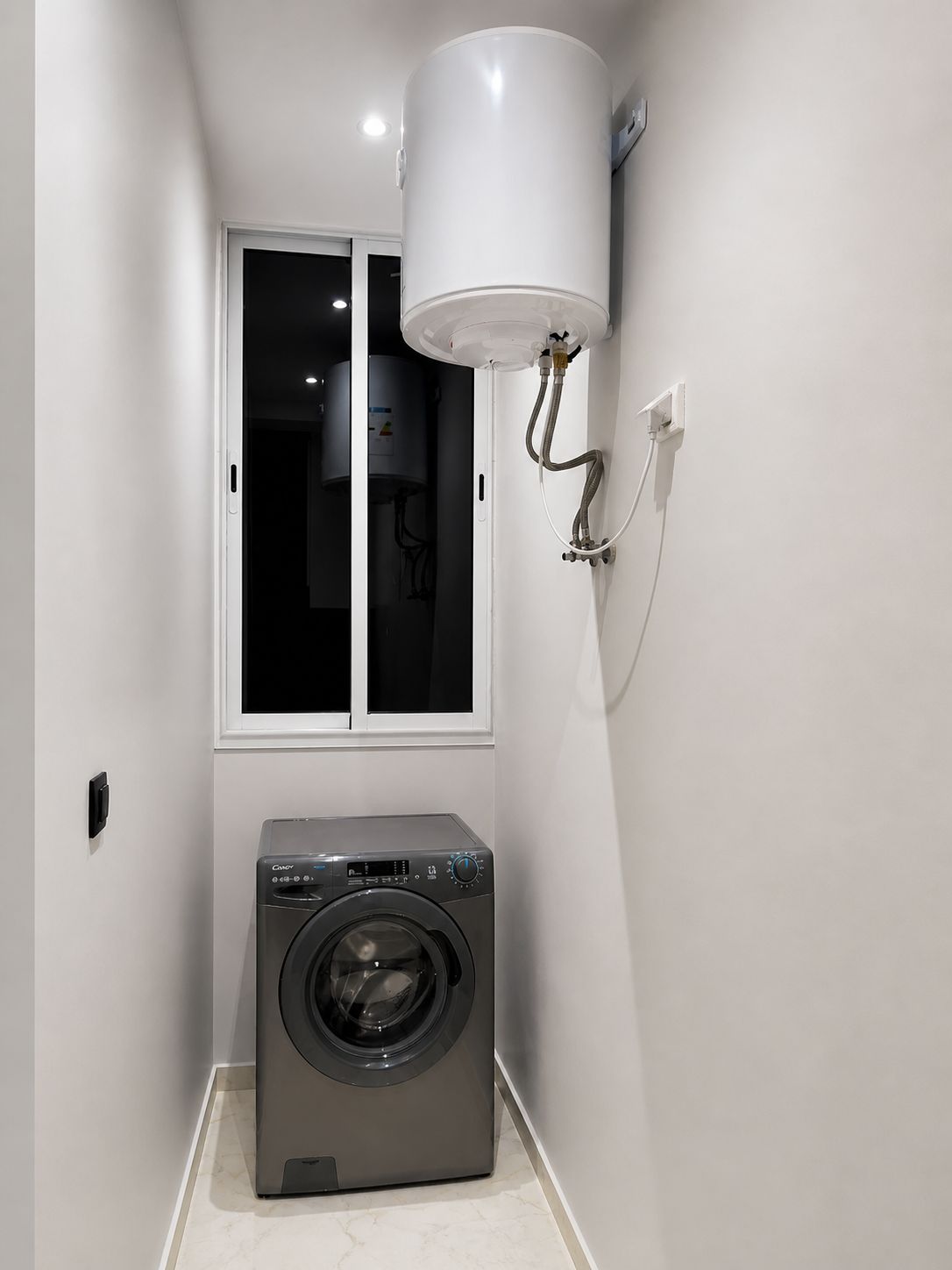 Ein kleiner Hauswirtschaftsraum mit einer Waschmaschine mit Frontlader, einem Fenster und einem wandmontierten Durchlauferhitzer.