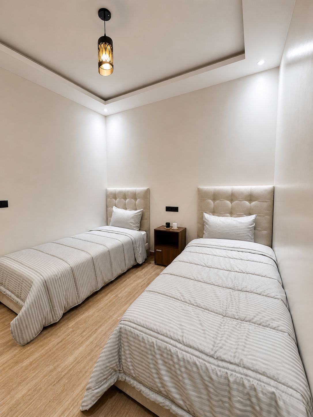 A bedroom with two beds, a nightstand, and a hanging light fixture.