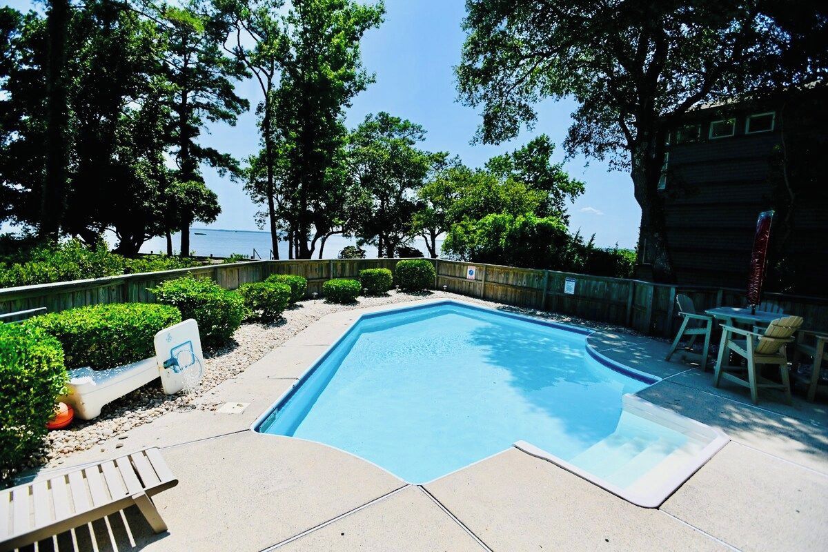 Una piscina con cubierta de madera, rodeada de árboles y vista al mar.