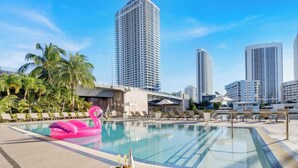 Ein Swimmingpool mit Liegestühlen und einem Flamingo-Schwimmpferd, umgeben von hohen Gebäuden und Palmen.