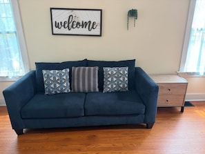 A blue sofa with patterned cushions, a wooden nightstand, a framed 'welcome' sign, and a hanging plant. - 3-bedroom house in tranquil Champaign with air conditioning (Champaign)