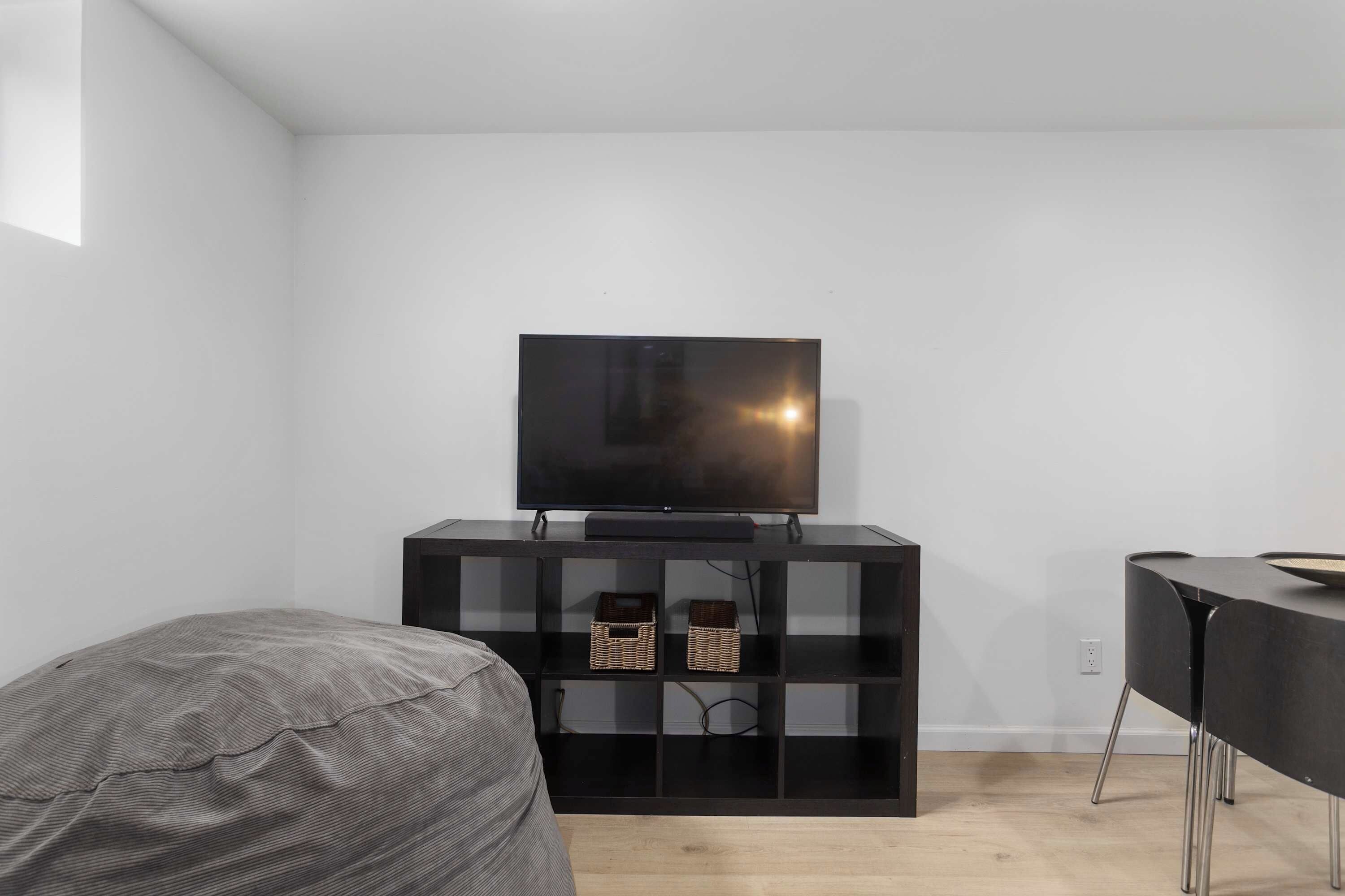 Una habitación moderna con un televisor de pantalla plana sobre un soporte negro, un sillón informal de color gris y una mesita con una cesta.