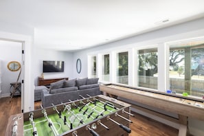 A living room with a foosball table, a grey sofa, a television, and a window with a view of the outdoors. - Coastal Home in Duck w/ Pool & All New Game Room! (Duck)