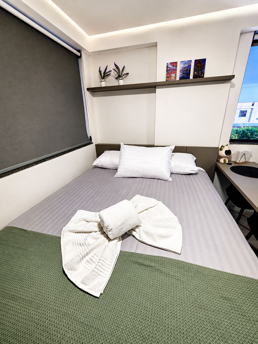 A hotel room with a bed, a desk, a chair, a window, and a wall-mounted shelf with decorative items.
