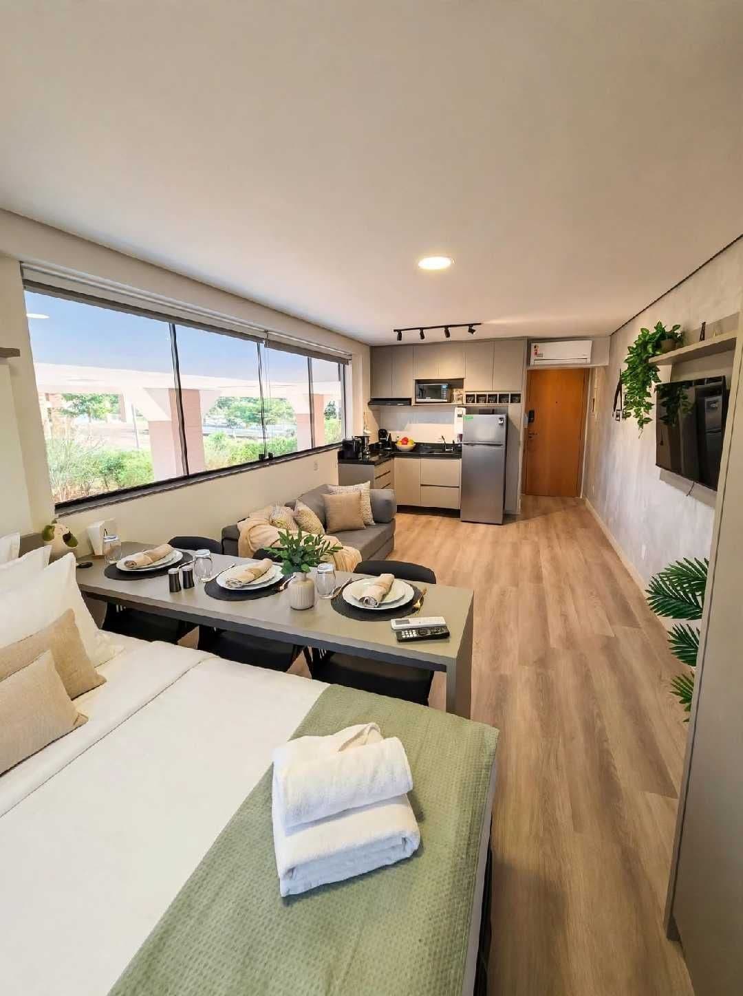 A modern hotel room with a dining area, kitchenette, and a large window providing a view of the outdoors.