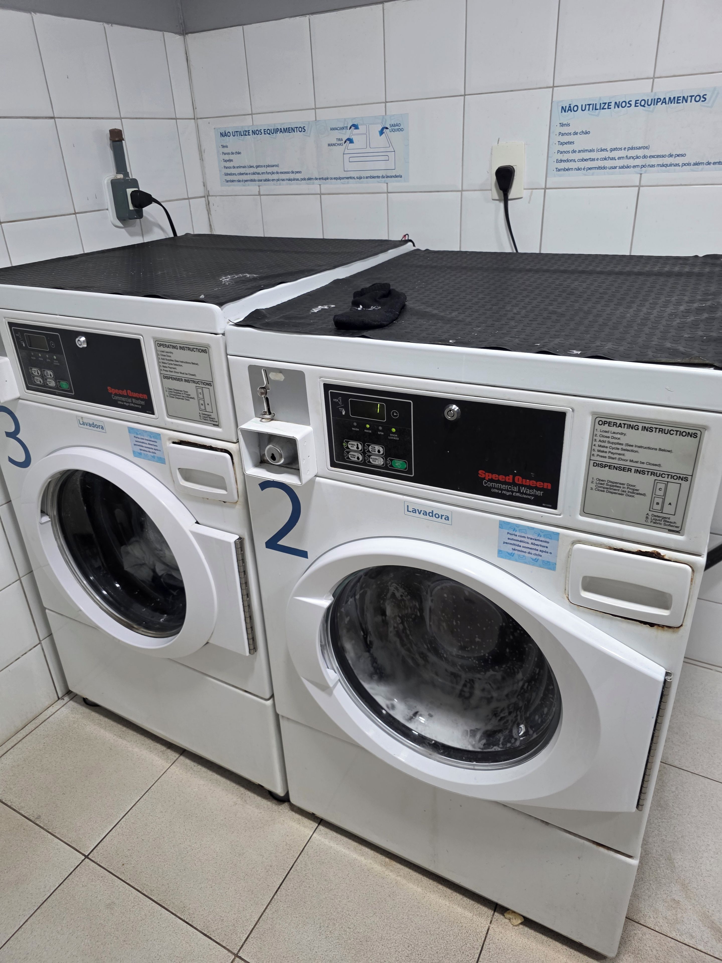 Two front-loading washing machines in a laundry area with operating instructions and safety warnings on the wall.
