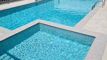 Una piscina rectangular con bordeado de azulejos, rodeada por una valla metálica, y una pequeña sección de un edificio con ventanas y una puerta al fondo.