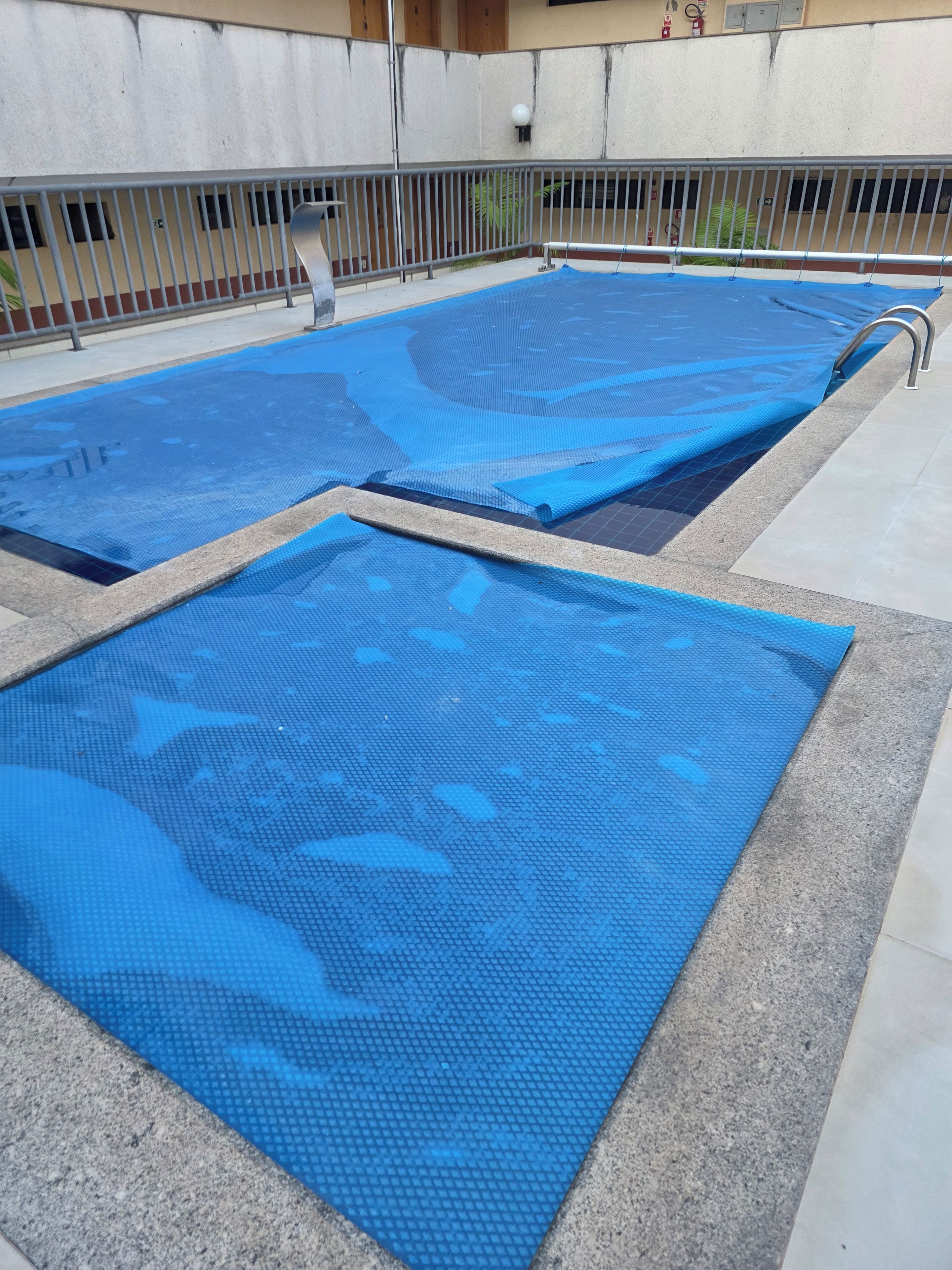 Piscina com tapetes azuis cobrindo a superfície, grade de metal e um edifício ao fundo.