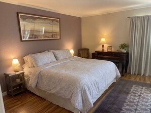 Una camera da letto con un letto, lampade da comodino, un comodino, una cassettiera, uno specchio, un quadro, una finestra con tende e un tappeto.