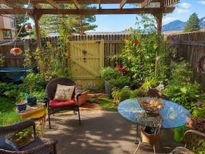 Ein Garten mit möbliertem Patio, einer Holz-Pergola, einem Korbstuhl mit Kissen, einem Glastisch mit dekorativer Schale und verschiedenen Pflanzen und Blumen.