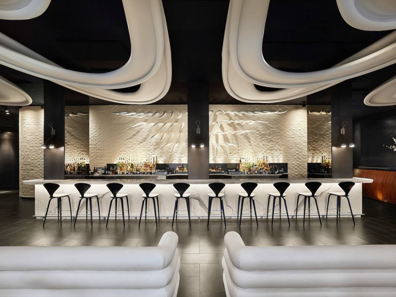 Una zona de bar con una barra larga, varios taburetes altos y una pared con textura de piedra.