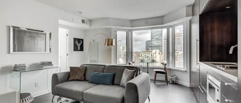 A modern apartment with a grey sofa, a glass coffee table, and a city view through large windows.