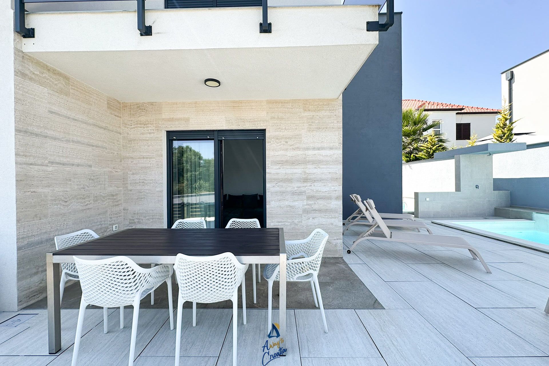 Um pátio externo com mesa e cadeiras, piscina e um edifício moderno ao fundo.